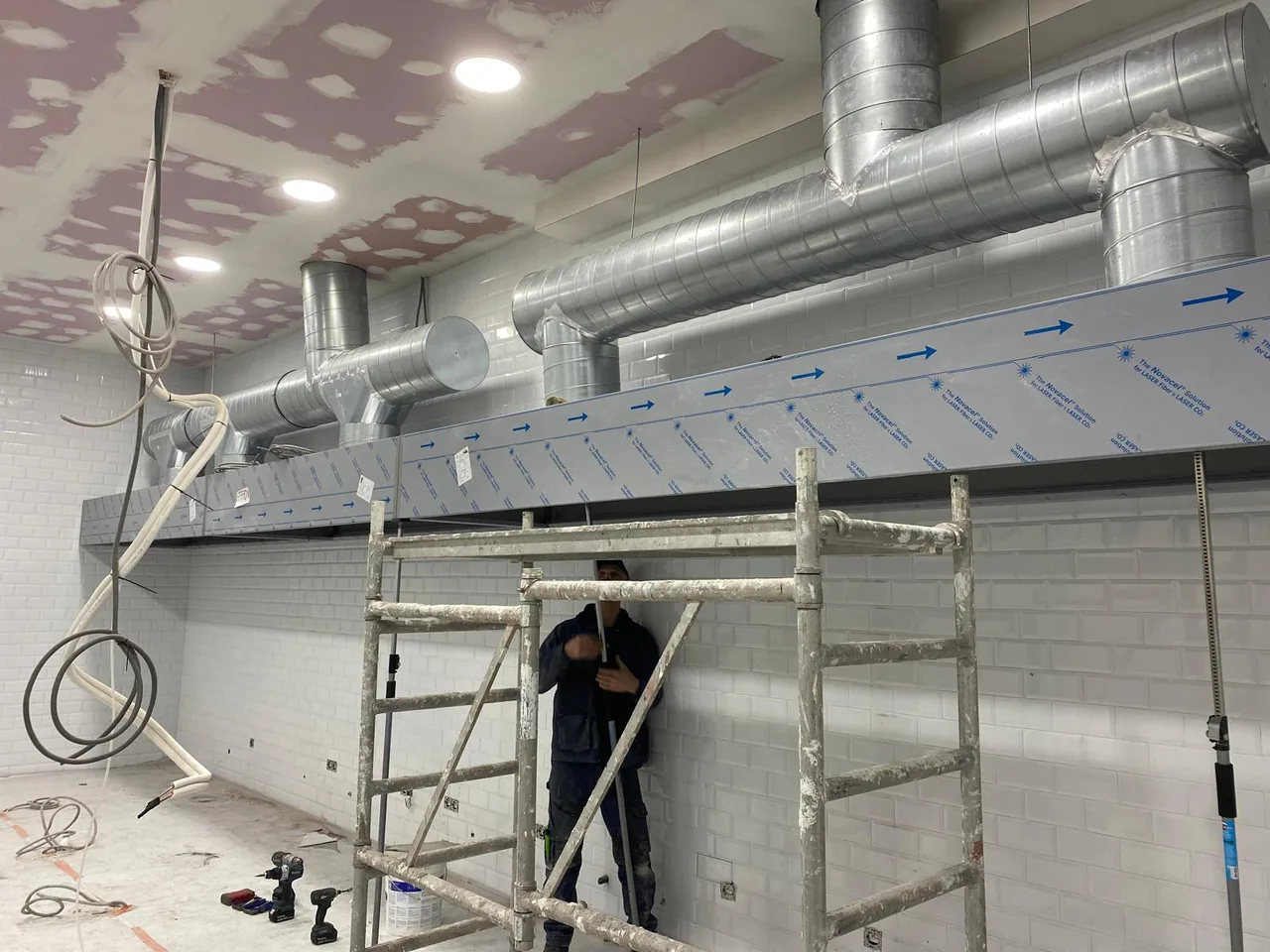 Technicien sur échafaudage installant un réseau de gaines spiralées galvanisées et un caisson d'extraction sur un chantier de cuisine industrielle